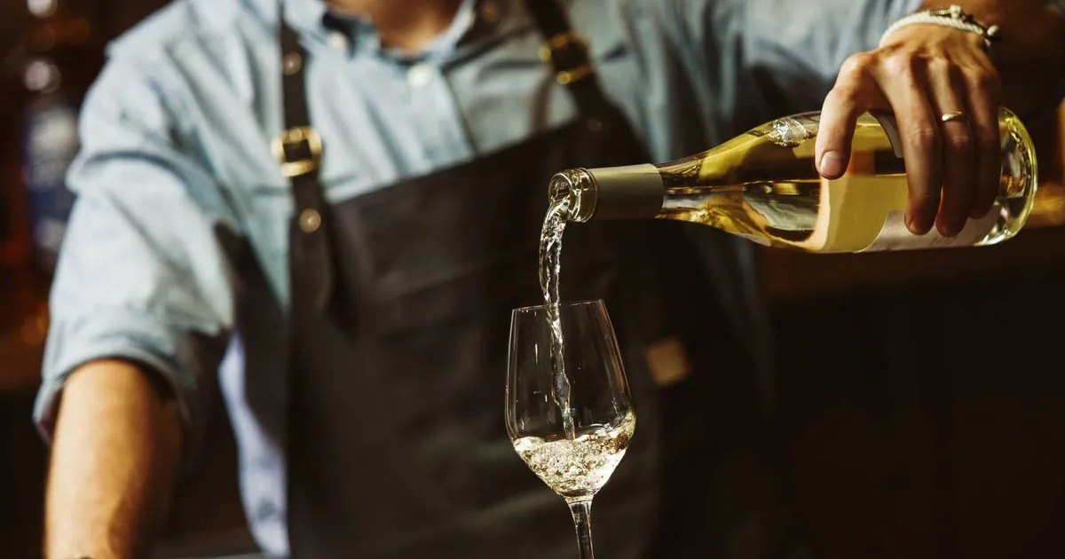 Un serveur en tablier verse du vin blanc dans un verre